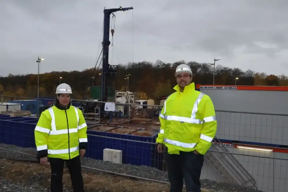 Zwei Mitarbeiter*innen der BGE vor einem Bohrplatz. Sie tragen gelbe Schutzwesten mit silbernen Reflektoren sowie weiße Bauhelme.