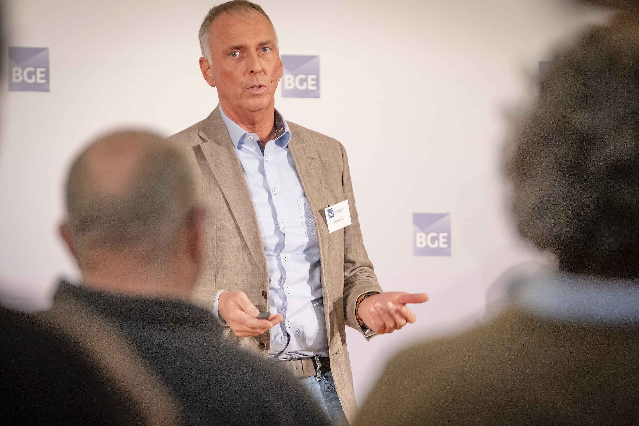 Portraitfoto eins Mannes mit grauem Haar, hellblauem Hemd und Sakko. Er spricht gestikulierend und trägt ein Namensschild an der Kleidung.