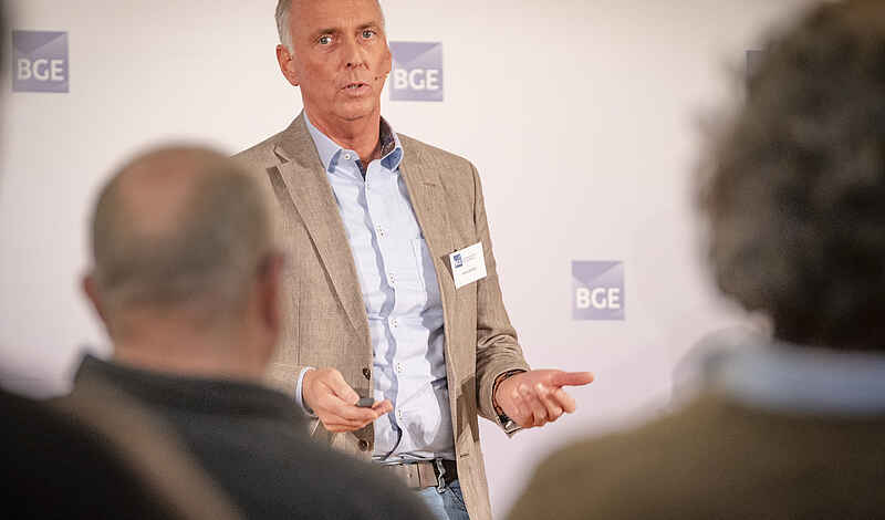 Portraitfoto eins Mannes mit grauem Haar, hellblauem Hemd und Sakko. Er spricht gestikulierend und trägt ein Namensschild an der Kleidung.