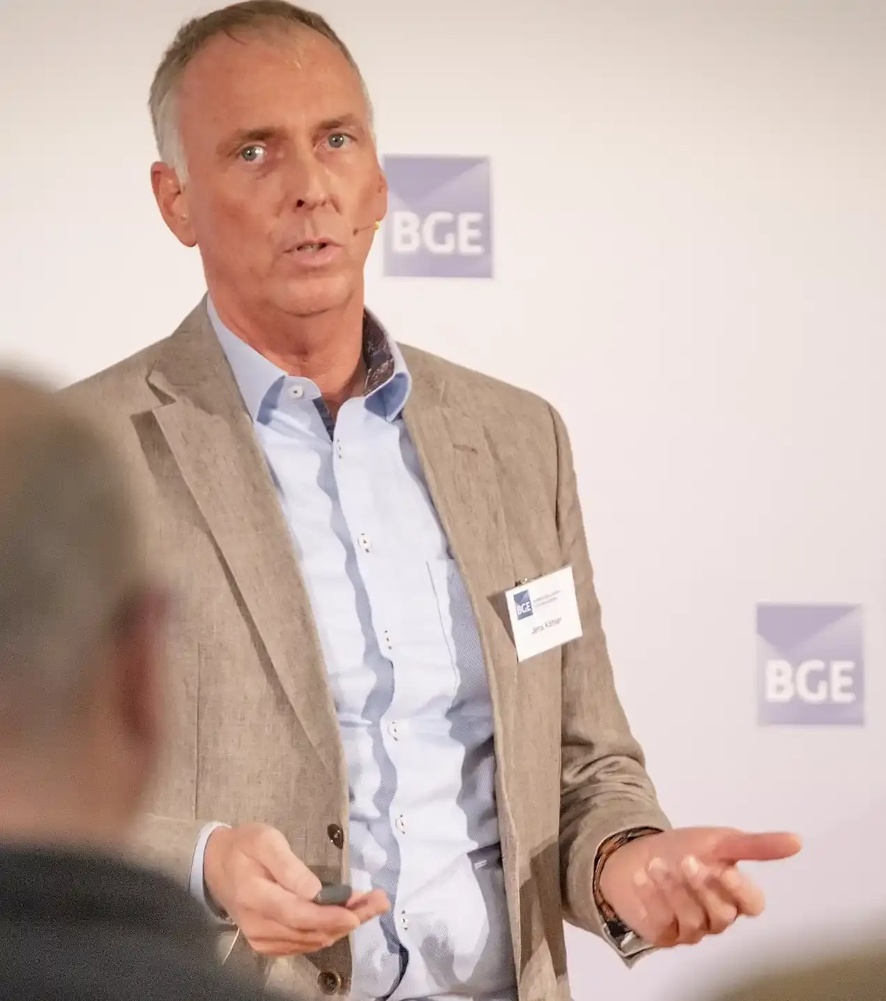 Portraitfoto eins Mannes mit grauem Haar, hellblauem Hemd und Sakko. Er spricht gestikulierend und trägt ein Namensschild an der Kleidung.