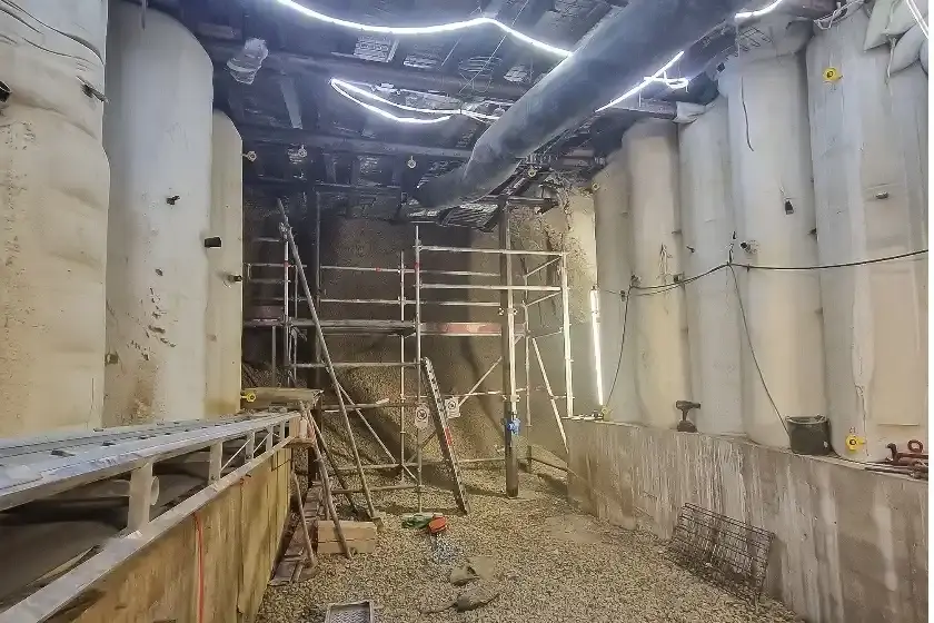 Das Bild zeigt den Blick in einen Abbau im Bergwerk. Auf der linken und rechten Seite stehen helle, säulenartige Stützbauwerke auf einem grauen Betonfundament. Quer über die Stützbauwerke, direkt unterhalb der Decke, verlaufen dunkle Metallträger. Zwischen den Stützbauwerken stehen Leitern und Gerüste. Im Hintergrund und auf dem Boden liegt Schotter