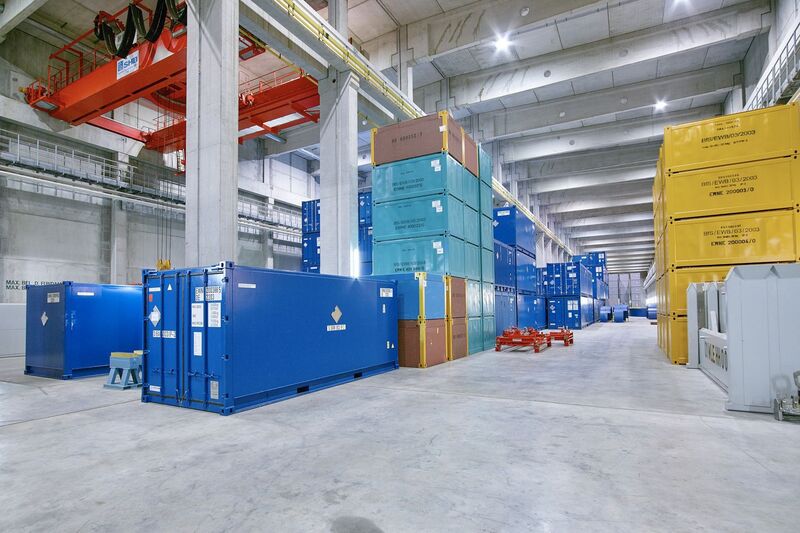 Container in verschiedenen Farben stehen teils gestapelt in einer großen Halle.