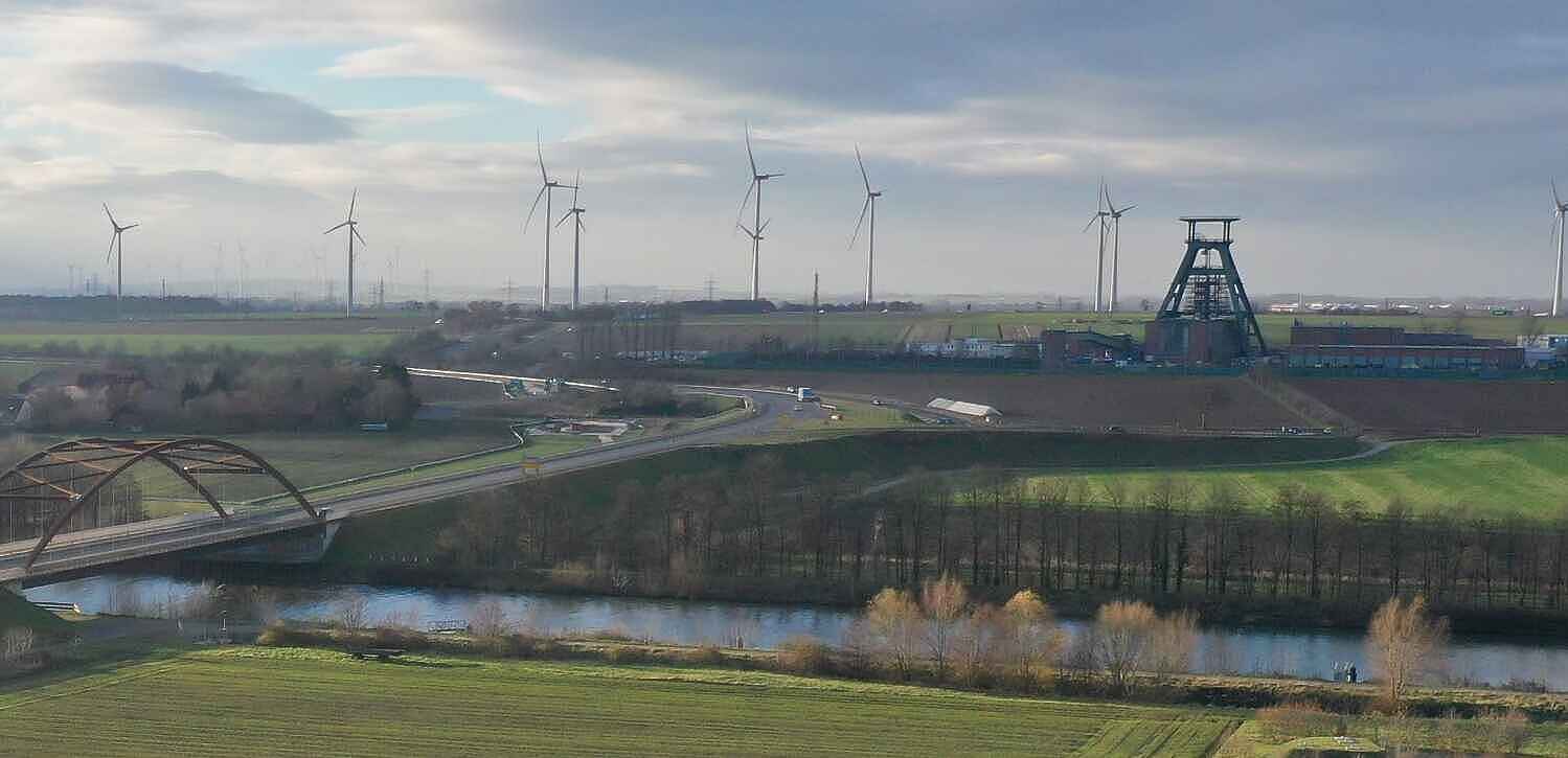 Luftaufnahme: Vor leicht bewölktem Himmel und zahlreichen Windrädern am Horizint ist die Schachtanlage 1 des Endlagers Konrad mit ihrem markanten Förderturm zu sehen. Im Vordergrund verläuft der Salzgitter Stichkanal. Darüber führt eine Autobrücke.