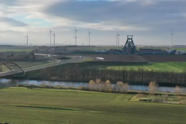 Luftaufnahme: Vor leicht bewölktem Himmel und zahlreichen Windrädern am Horizint ist die Schachtanlage 1 des Endlagers Konrad mit ihrem markanten Förderturm zu sehen. Im Vordergrund verläuft der Salzgitter Stichkanal. Darüber führt eine Autobrücke.