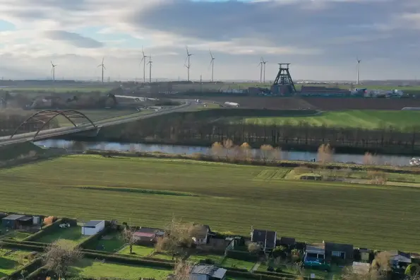 Aerial photograph: backed by a slightly cloudy sky with numerous wind turbines on the horizon, you can see Shaft 1 of the Konrad repository with its striking winding tower. The Salzgitter Stichkanal passes in the foreground with a motorway bridge running over it.