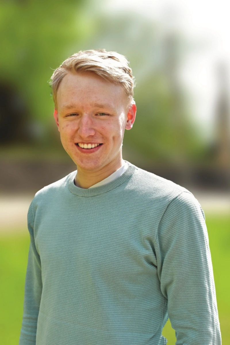 Portrait eines jungen Mannes mit rot-blonden kurzen Haaren in einem türkisfarbenen Pullover.