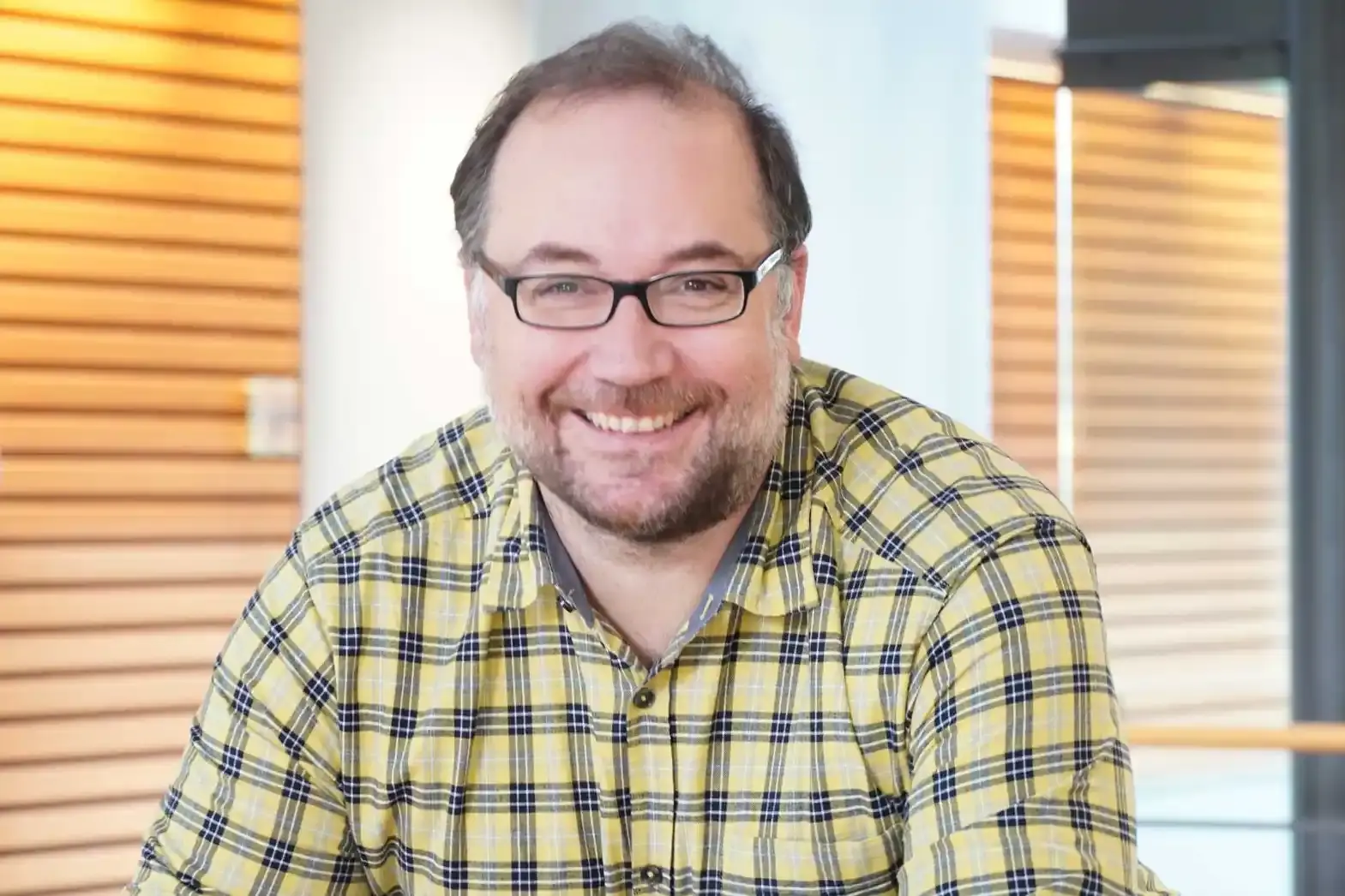 Porträtbild: Ein Mann mit dunklen kurzen Haare, Brille und 3-Tage-Bart lächelt in die Kamera. Er trägt ein kariertes Hemd. 