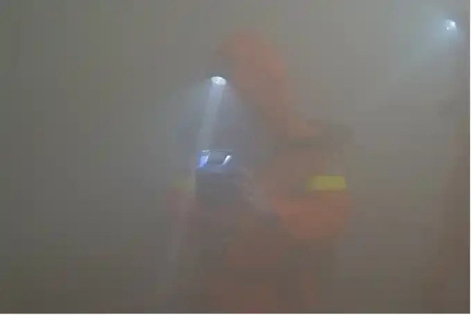 Eine Person in einem orangefarbenen Rettungsanzug und Helmlampe steht in dichtem Rauch. Der Lichtkegel der Helmlampe fällt auf einen Gegenstand, welche die Person in ihren Händen hält.