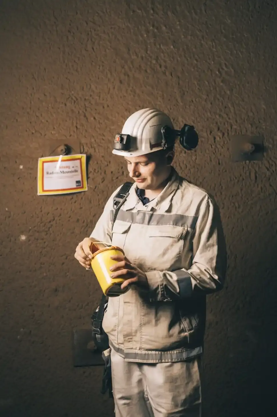 Ein Mann unter Tage in weißer Arbeitskleidung und weißem Helm hält eine gelbe Dose in den Händen, die er gerade öffnet.