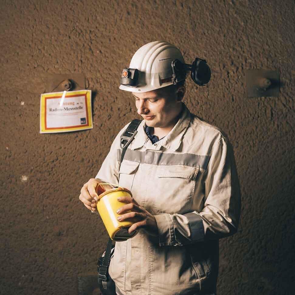 Ein Mann unter Tage in weißer Arbeitskleidung und weißem Helm hält eine gelbe Dose in den Händen, die er gerade öffnet.