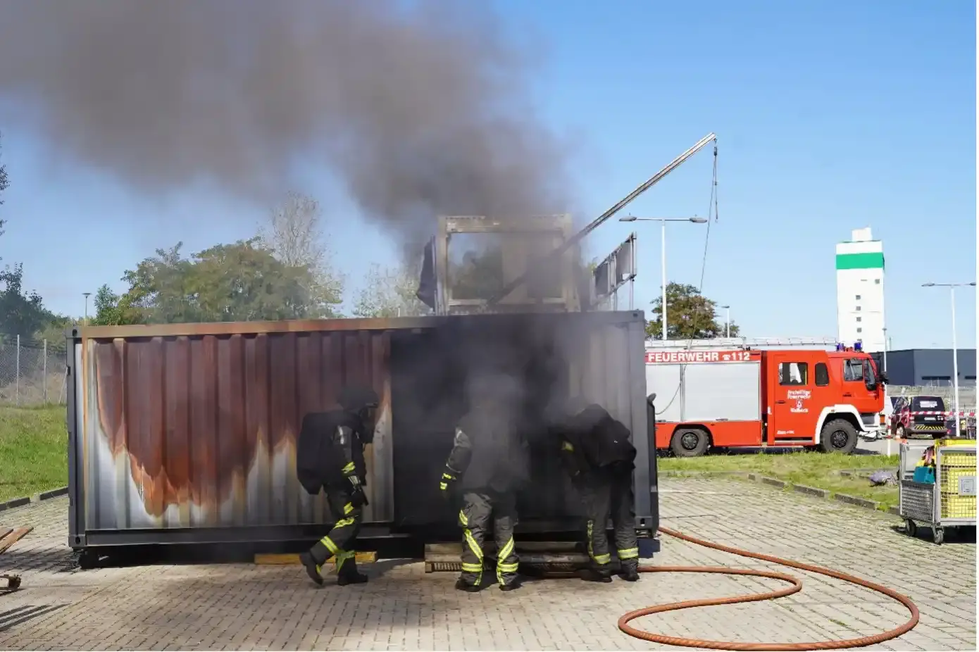Drei Personen in Schutzausrüstung betreten einen qualmenden Container. Im Hintergrund stehen ein Löschfahrzeug und der hohe weiße Förderturm Bartensleben. 