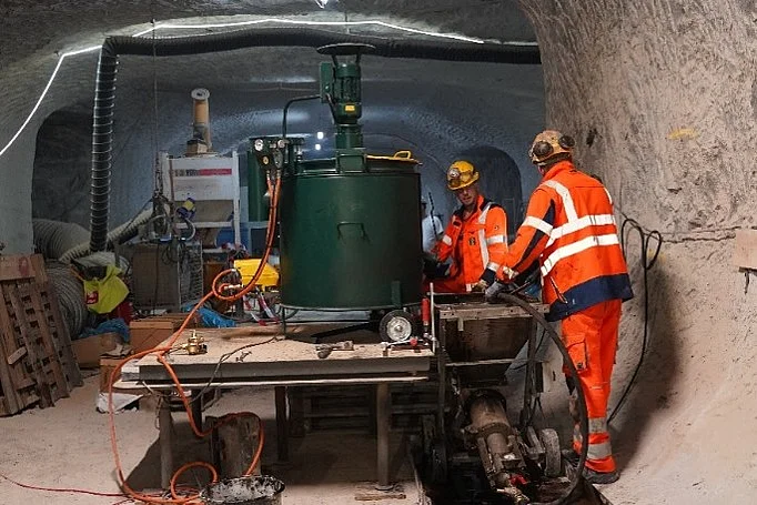 In einem Raum unter Tage steht ein großer dunkelgrüner Kessel auf einem Tisch. Neben dem Tisch stehen zwei Bergleute. Sie tragen Schutzhelm und Arbeitskleidung. 