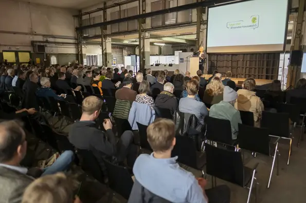Foto eines Veranstaltungsraumes. Viele Personen sitzen mit dem Rücken zur Kamera gewandt auf dunklen Stühlen und blicken in Richtung Podium. Auf deisem spricht eine Frau mit blonden, schulterlangen Haaren zu den Anwesenden.