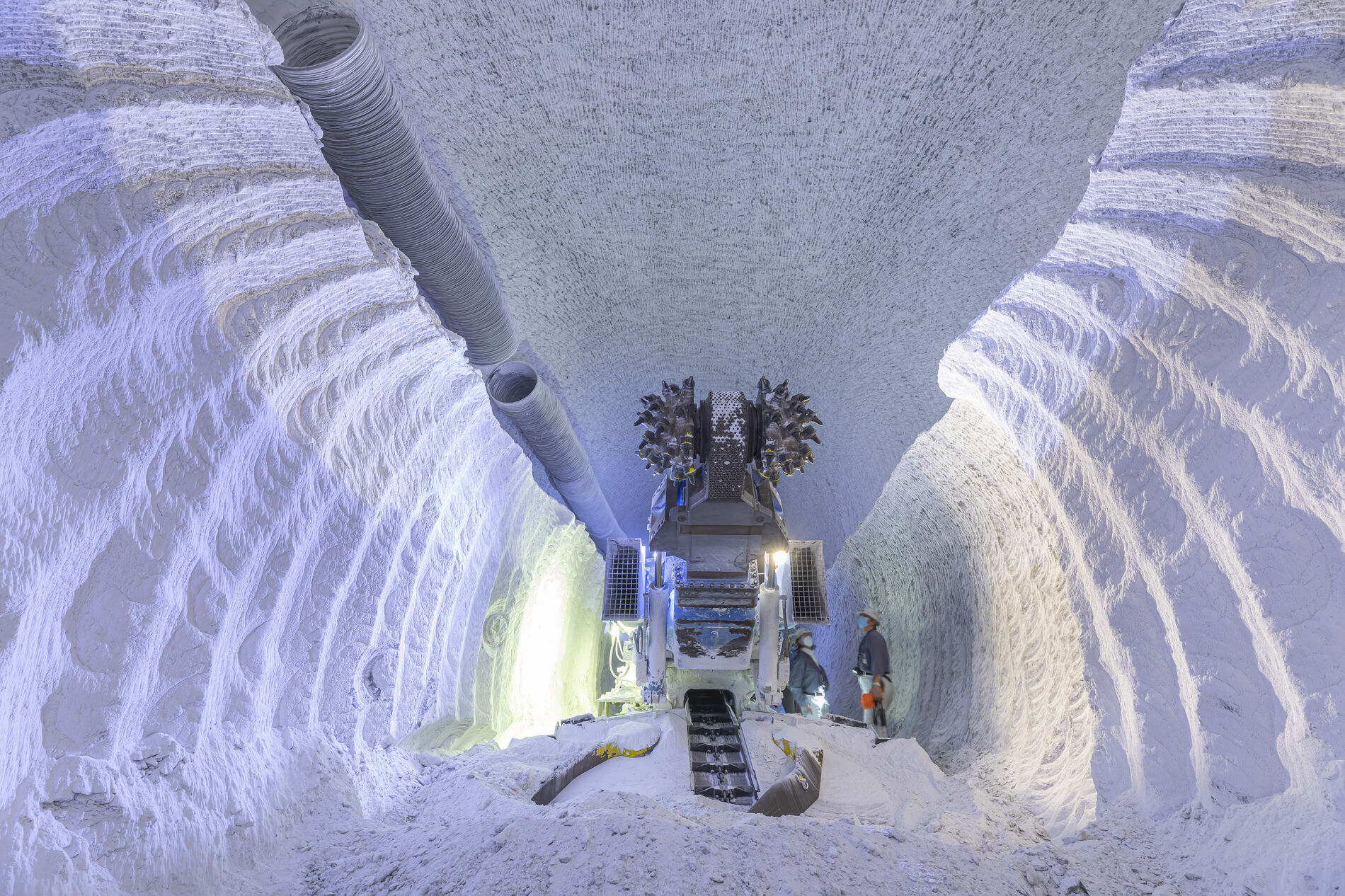 Two miners stand next to a large machine in a white chamber made of salt underground.