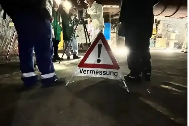 Eine Person in weißer Schutzkleidung steht an einem dreibeinigen Stativ. Im Vordergrund steht ein dreieckiges Schild mit der Aufschrift „Vermessung“. 