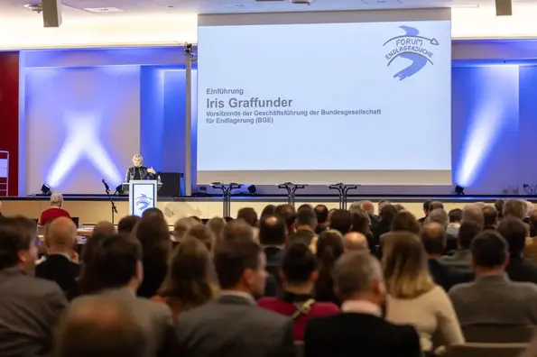 Über mehrere gut besetzte Stuhlreihen hinweg wurde eine Bühne fotografiert, auf der eine Frau an einem Redner*innenpult steht. Sie hat blonde kinnlange Haare, gestikuliert und trägt eine schwarze Lederjacke.