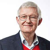 Porträtbild: Ein Mann mit hellgrauen kurzen Haaren und Brille. Unter einem schwarzen Sakko trägt er ein helles Hemd und einen roten Pullunder.