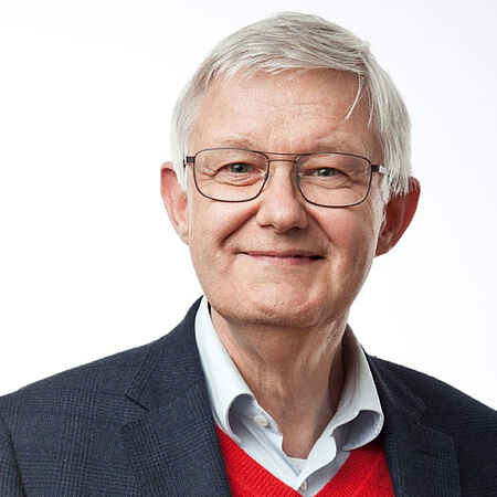 Porträtbild: Ein Mann mit hellgrauen kurzen Haaren und Brille. Unter einem schwarzen Sakko trägt er ein helles Hemd und einen roten Pullunder.