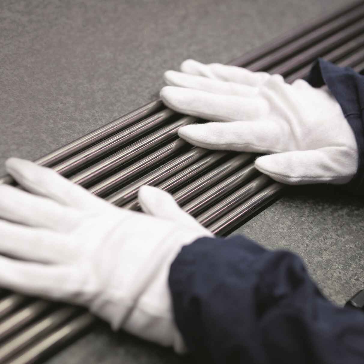 Zwei Hände in weißen Handschuhen liegen auf mehreren dicht beieinanderliegenden metallisch glänzenden Stäben.