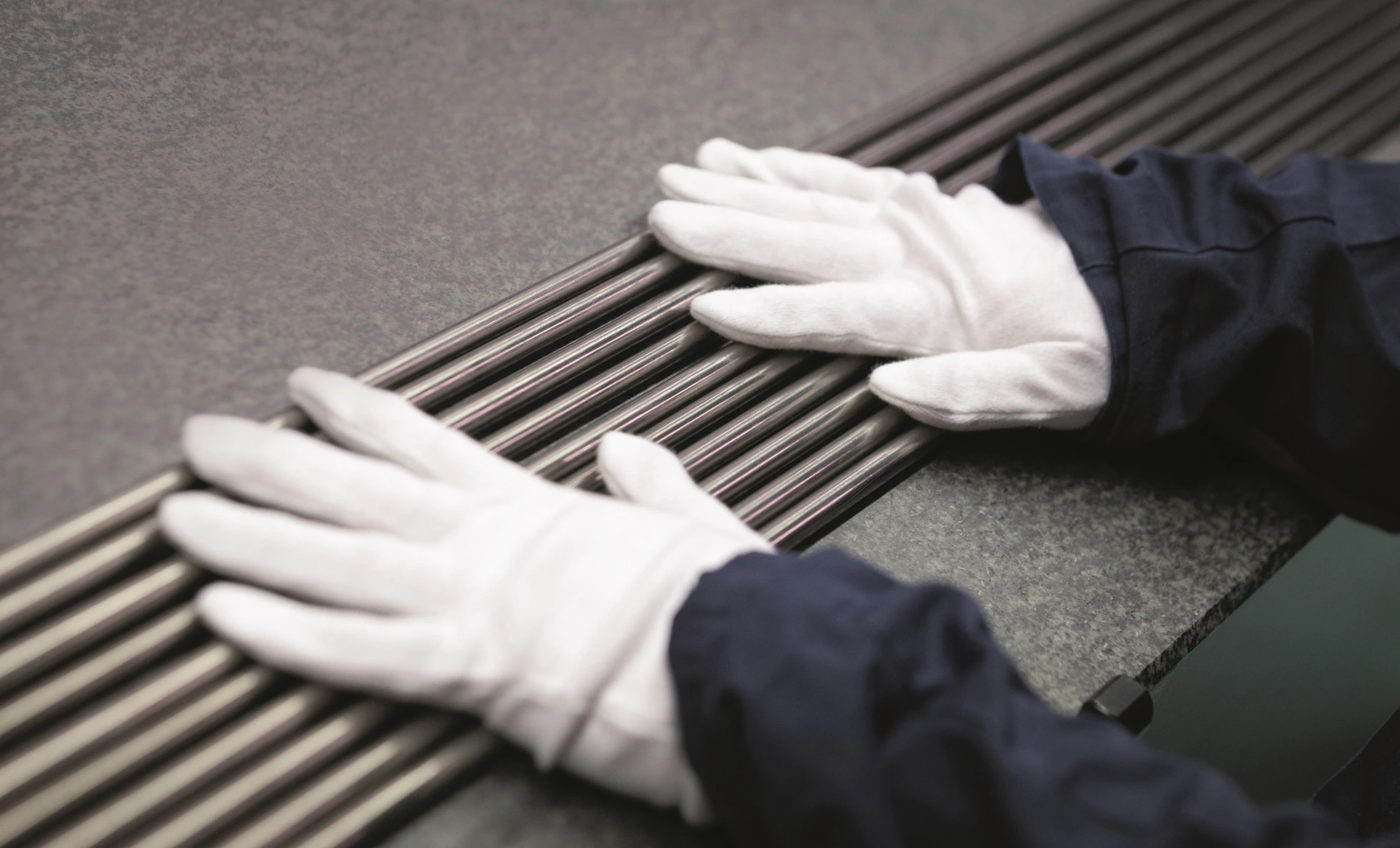 Zwei Hände in weißen Handschuhen liegen auf mehreren dicht beieinanderliegenden metallisch glänzenden Stäben.