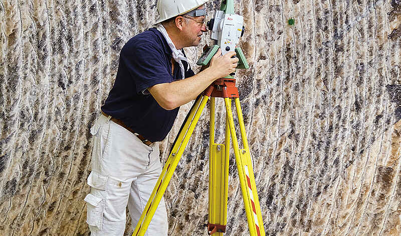 Vermessungsingenieur mit gelbem Schutzhelm richtet im Untergrund ein Theodolit-Messgerät auf einem Stativ aus.