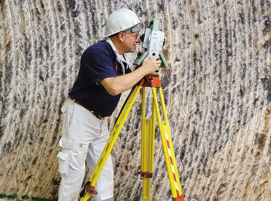 Vermessungsingenieur mit gelbem Schutzhelm richtet im Untergrund ein Theodolit-Messgerät auf einem Stativ aus.