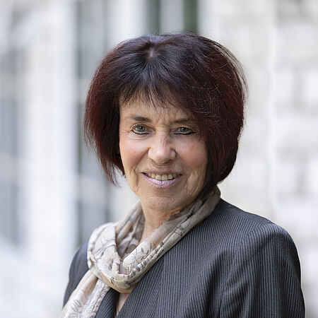 Eine Frau mit kinnlangen braunen Haaren lächelt freundlich in die Kamera. Sie trägt einen grauen Blazer und einen hellen gemusterten Schal. 