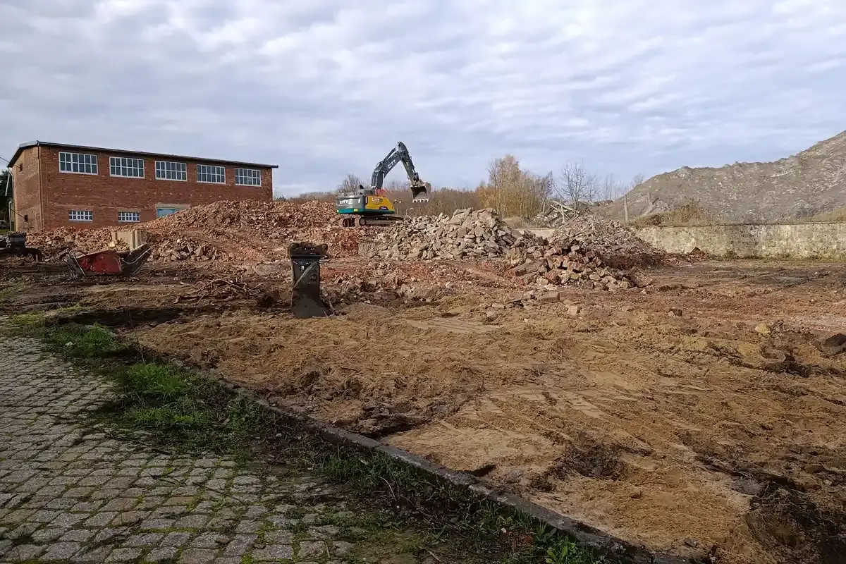 Ein Abrissbagger steht auf einer Halde aus Bauschutt vor einem bewölkten Himmel.