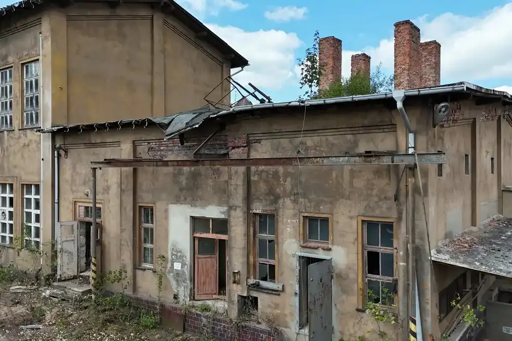 Ein altes Gebäude mit offenen Türen. Das Gebäude ist stark verfallen. Aus einem der Räume wächst ein Baum durch das Dach.