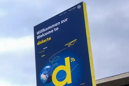 Rund 20 Personen befinden sich vor einem gläsernen Gebäude, vor dem riesige Aufsteller mit der Aufschrift "didacta" stehen. Einige bewegen sich auf das Gebäude zu. 