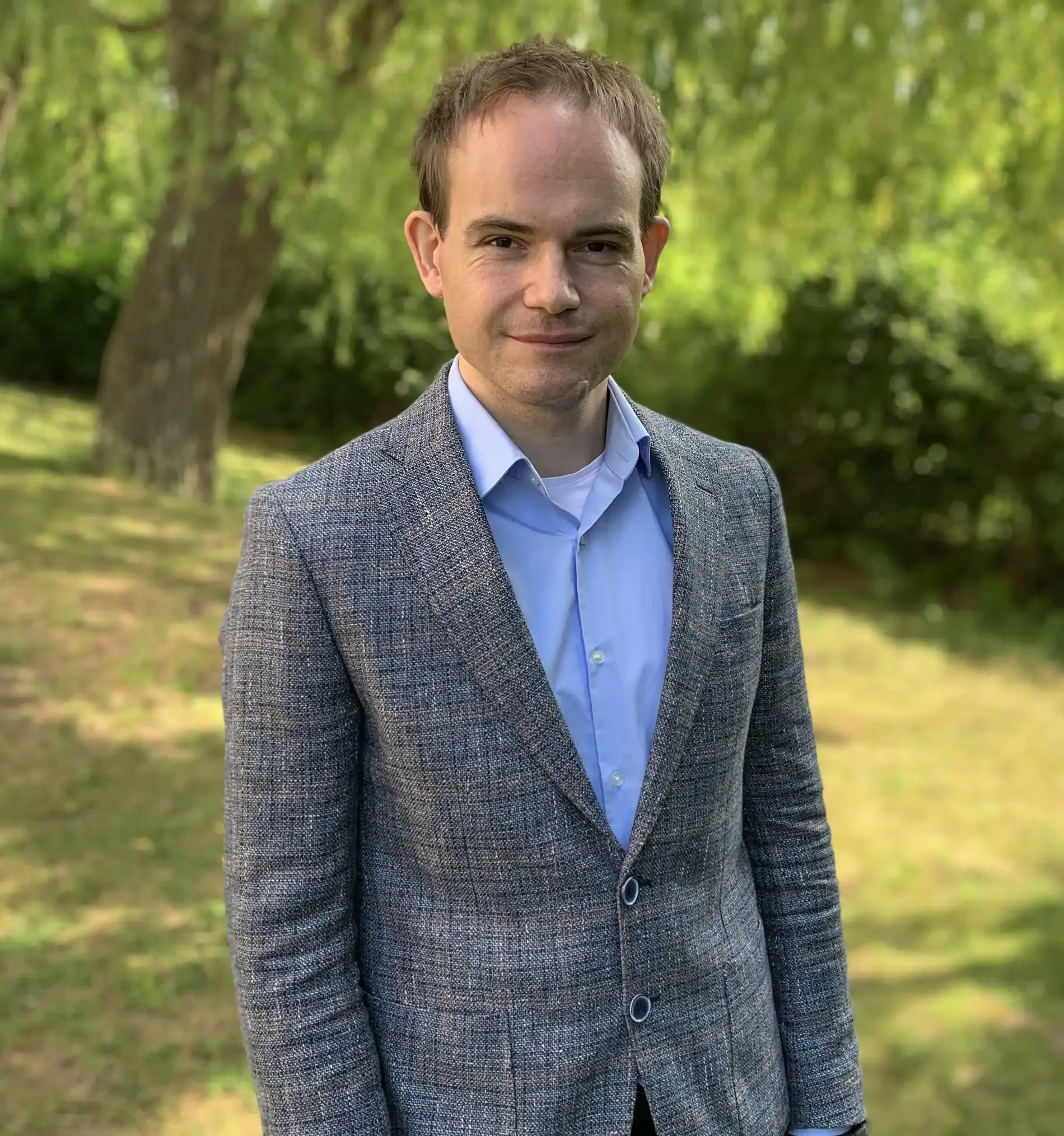 Ein Mann mit einem grauen Sakko und einem hellblauen Hemd lächelt in die Kamera. Er steht bei sommerlichem Wetter auf einer Wiese vor einer Weide.