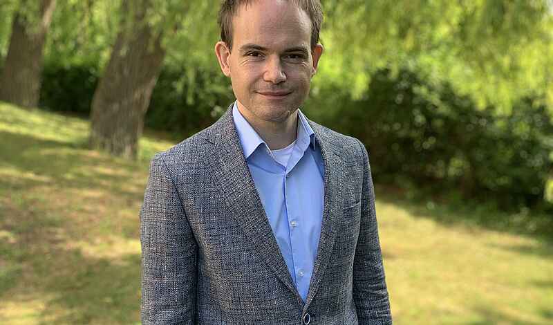 Ein Mann mit einem grauen Sakko und einem hellblauen Hemd lächelt in die Kamera. Er steht bei sommerlichem Wetter auf einer Wiese vor einer Weide.