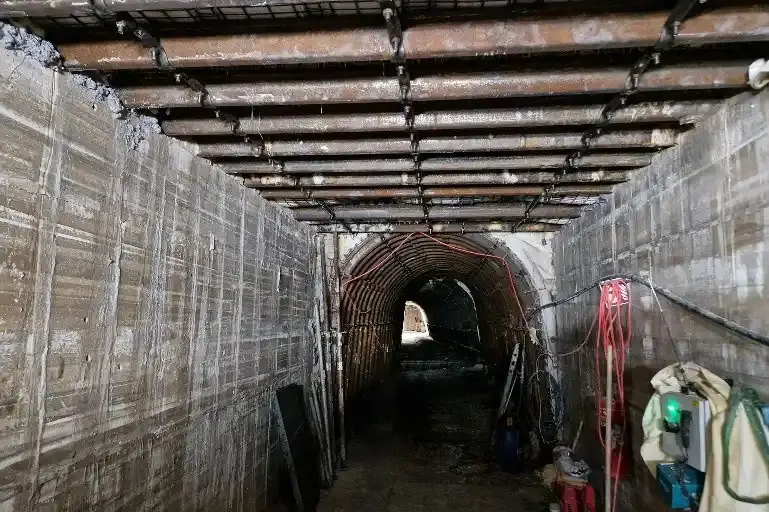 Das Bild zeigt den Blick in eine Strecke im Bergwerk. Über die ersten Meter erstrecken sich graue Betonwände an beiden Seiten der Strecke. Quer auf den Betonwänden sind mehrere Stahlträger verbaut, die die Wände verbinden. Im hinteren Teil schließt sich ein Bereich an, in dem die Strecke durch mehrere, hintereinander verbaute, halbrunde Stahlbögen gestützt wird.