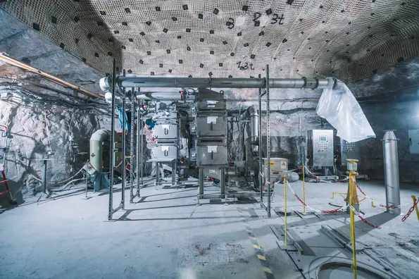 Unterirdischer Raum mit grob behauenen Felswänden und Decke. In der Mitte steht eine große technische Anlage aus Metallrohren, Tanks und Steuerungselementen, umgeben von Absperrungen und Warnmarkierungen. Kabel und Lüftungsrohre verlaufen entlang der Decke und Wände.