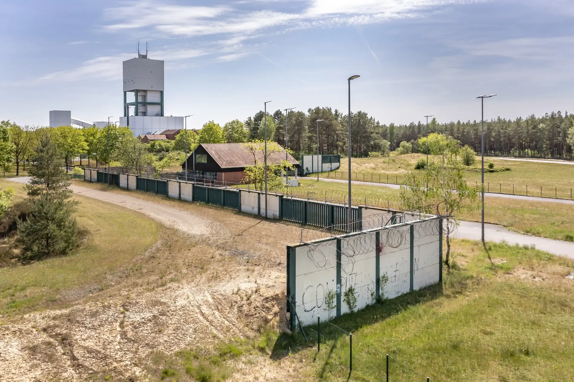 The remains of a white wall can be seen in the foreground. In the background, the winding tower of the Gorleben mine rises up behind trees. 