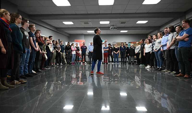 Auf dem Foto ist eine Person zu sehen, die in der Mitte eines Halbkreises von Jugendlichen und jungen Erwachsenen steht und ihnen etwas vorträgt.