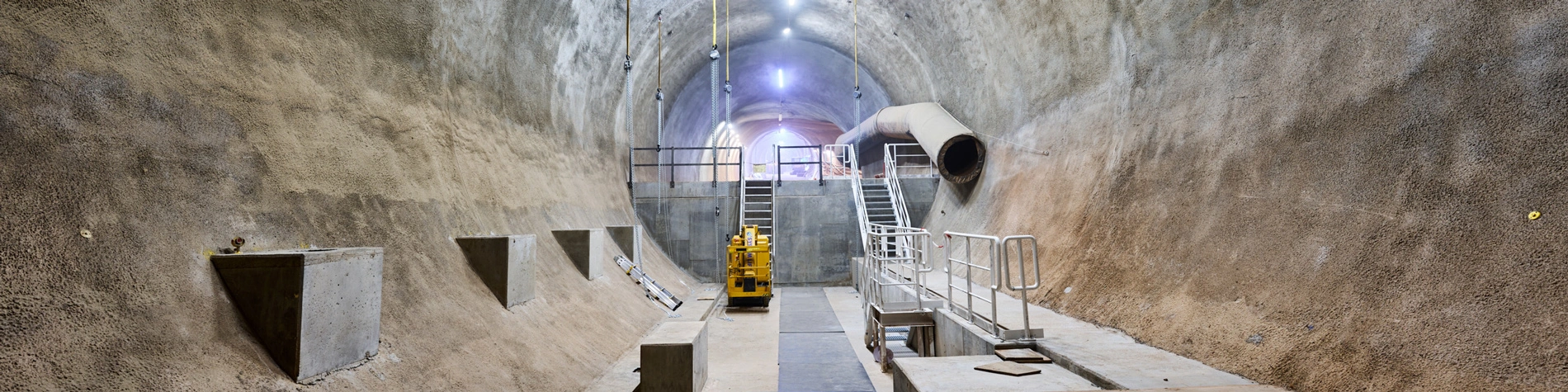 Auf dem Foto ist eine runde Strecke in einem Bergwerk zu sehen. Ein gelbes Gerät steht mittig der Strecke und zwei Treppen sind erkennbar, die auf eine höhere Ebene der Strecke führen.