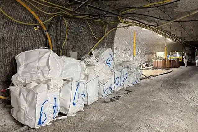 Foto von großen, befüllten weißen Kunststoffsäcken mit blauen Aufschriften unter Tage in einem Salzbergwerk. Im hinteren Bildteil gehen Personen in weißer Arbeitskleidung.