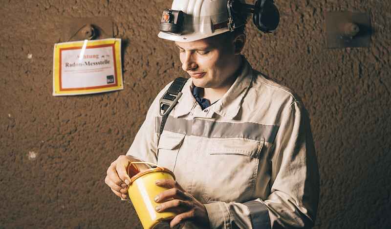 Ein Mann unter Tage in weißer Arbeitskleidung und weißem Helm hält eine gelbe Dose in den Händen, die er gerade öffnet.