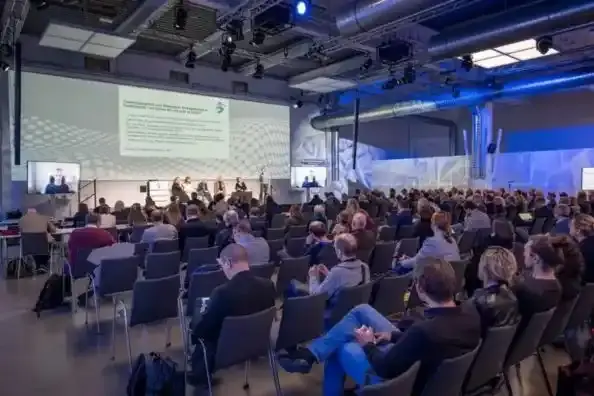 Großer Konferenzraum mit vielen Teilnehmenden, die einer Präsentation auf einer Leinwand folgen.