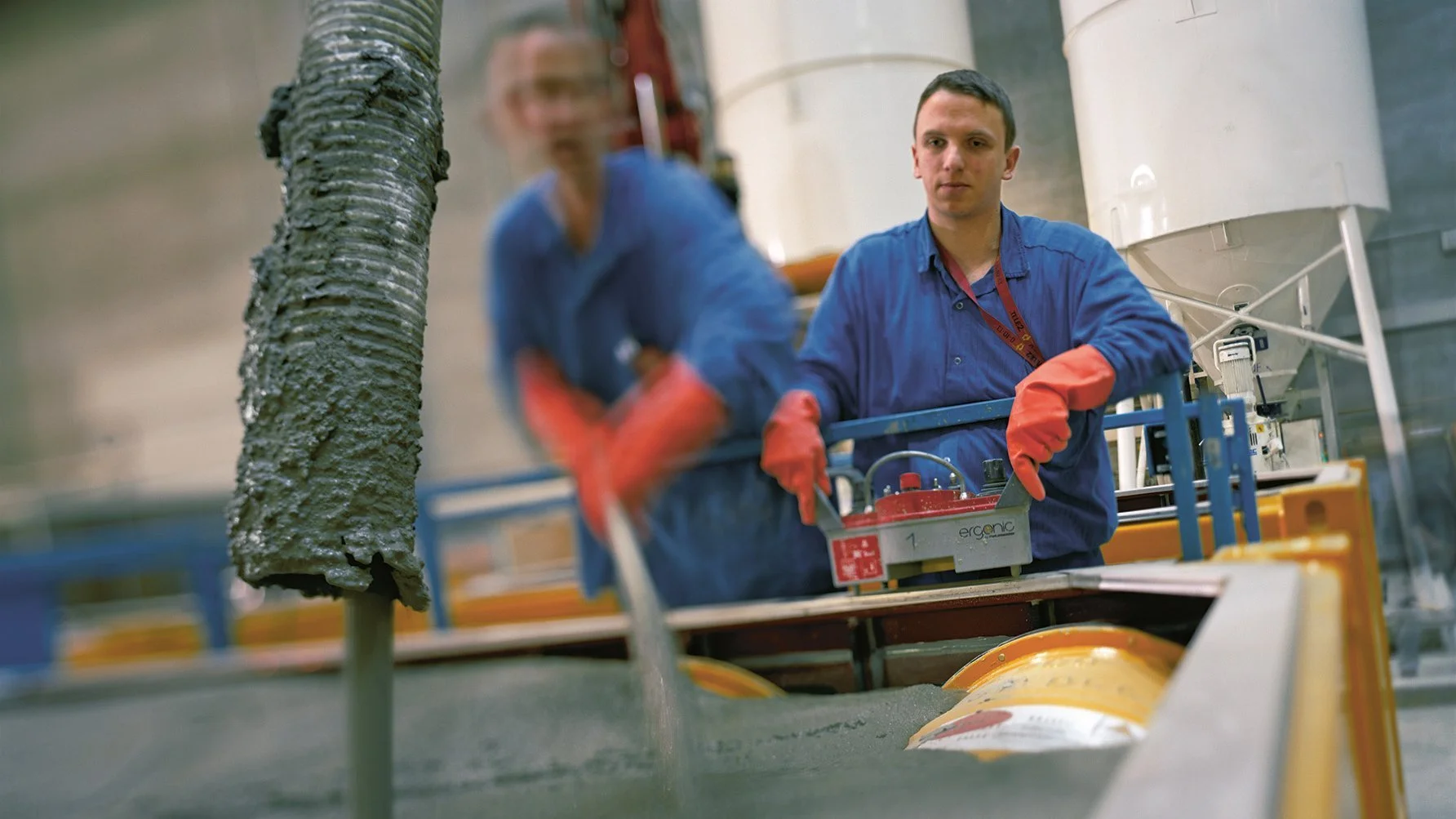 Zwei Männer in blauer Arbeitskleidung mit Fernbedienung. Aus einem Rohr kommt Beton heraus.