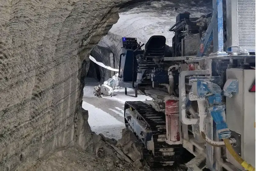 Auf der rechten Seite des Bildes ist im Anschnitt eine Fräsmaschine mit Kettenantrieb abgebildet. Diese steht im Zugangsbereich zu einem Streckenbereich im Bergwerk. Der Fräskopf und Auslieger ragen in den Streckenbereich hinein. Auf der linken Seite ist eine Wand aus Salzgestein mit Frässpuren zu erkennen.