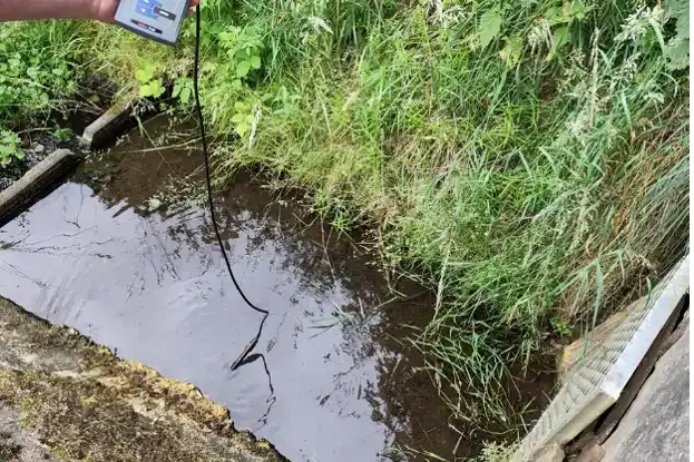 Blick auf ein Gewässer, die Uferböschung ist auf der unteren Seite gemauert, oben ist die Böschung begrünt. Eine Hand hält einen Wasserstandsanzeiger.