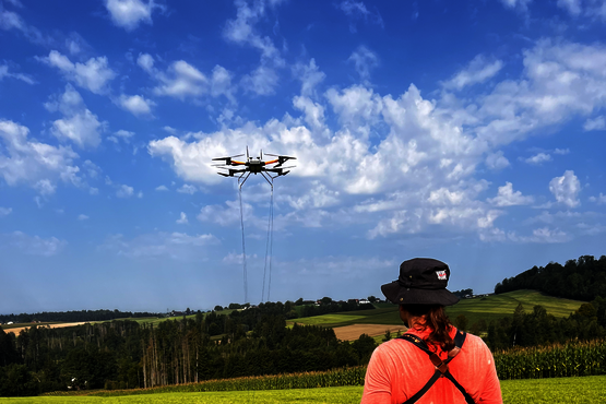 Das Foto zeigt eine Peron mit langen Haaren und Hut von hinten. Die Person steuert eine aufsteigende Drohne über ein Feld bei blauem Himmel.