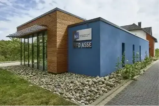 Zu sehen ist ein Gebäude. Ein Teil des Gebäudes ist blau angestrichen, ein anderer Teil mit Holz verschalt. Eine Fensterfront ermöglicht den Blick nach innen. Auf einem Schild steht Info Asse.