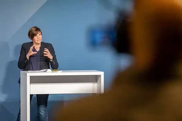 Eine Frau mit gescheitelten braunen Haaren, schwarzer Bluse und blau-weiß gepunktetem Hemd steht an einem weißen Rednerpult und hält einen Vortrag.