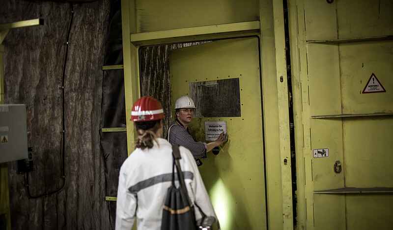 Zwei Frauen in Bergmannskluft stehen unter Tage vor einer massiven gelben Wettertür. Eine Frau öffnet die gelbe Tür einen Spalt weit und blickt in die Kamera. Die andere Frau steht mit dem Rücken zur Kamera. Sie trägt einen Rucksack.