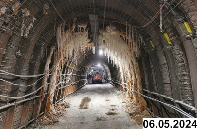 Unter Tage wird eine Strecke durch künstliches Licht erleuchtet. Die Strecke hat eine gerundete Decke, die durch Metallstützen gesichert wird. Am Ende der Strecke steht ein Fahrzeug. An der Decke haben sich viele Tropfsteine gebildet. Auf dem Boden sind Pfützen zu erkennen.