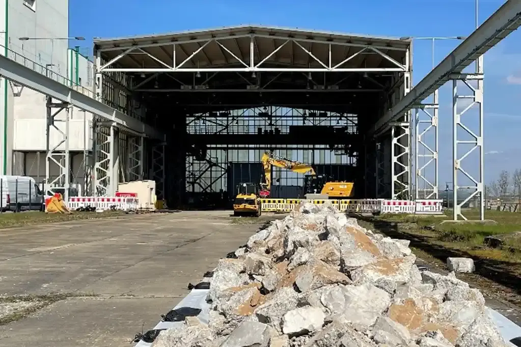 Foto eines Haufens von Bauschutt vor einer großen Industriehalle. In der Halle stehen Baumaschinen.