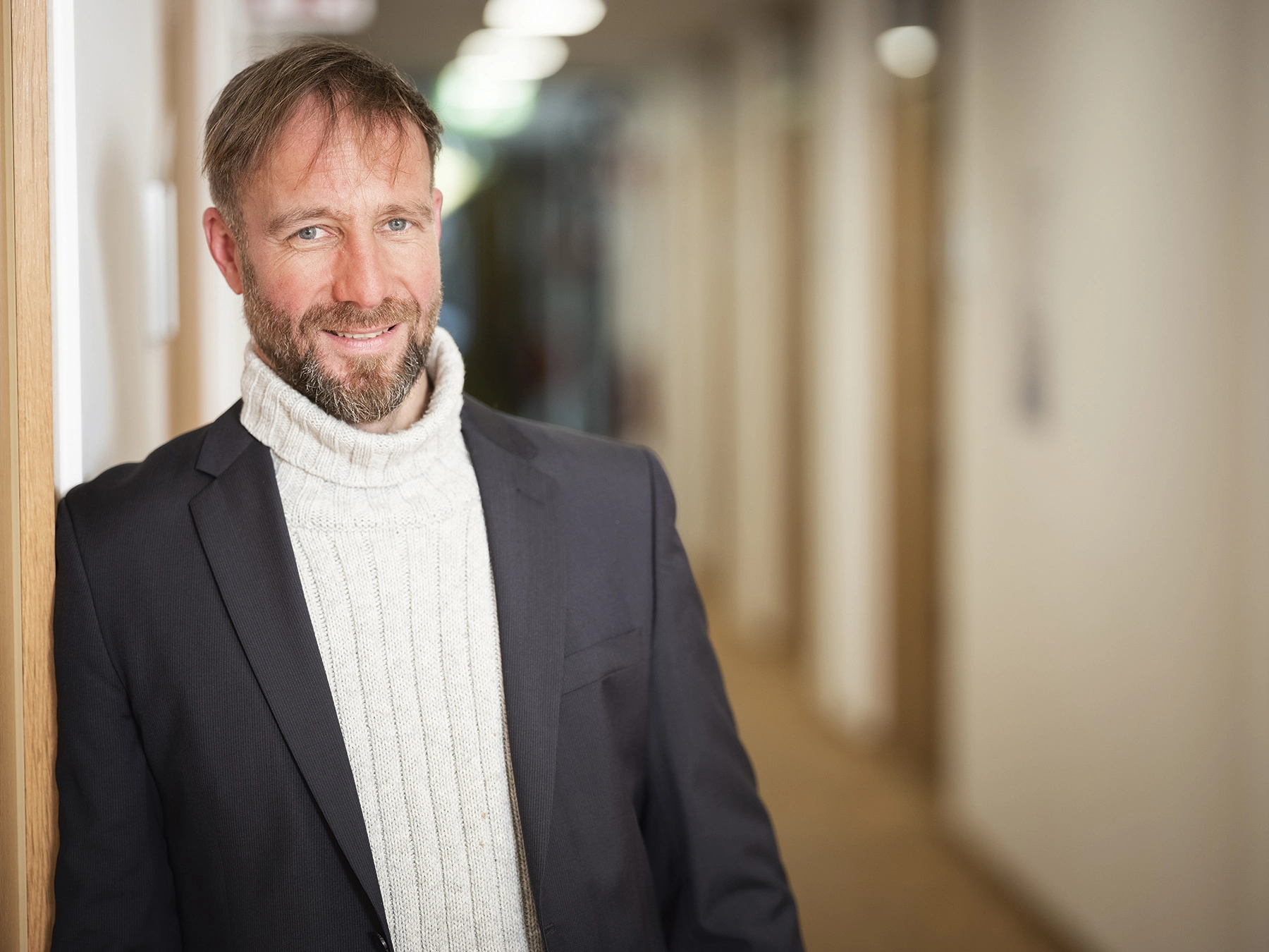 Ein Mann mit kurzen braunen Haaren und Bart lehnt lächelnd seitlich an einer Wand. Er trägt einen weißen Rollkragenpullover und ein schwarzes Sakko. Im Hintergrund ist unscharf ein Flur zu erkennen, der in die Tiefe führt.